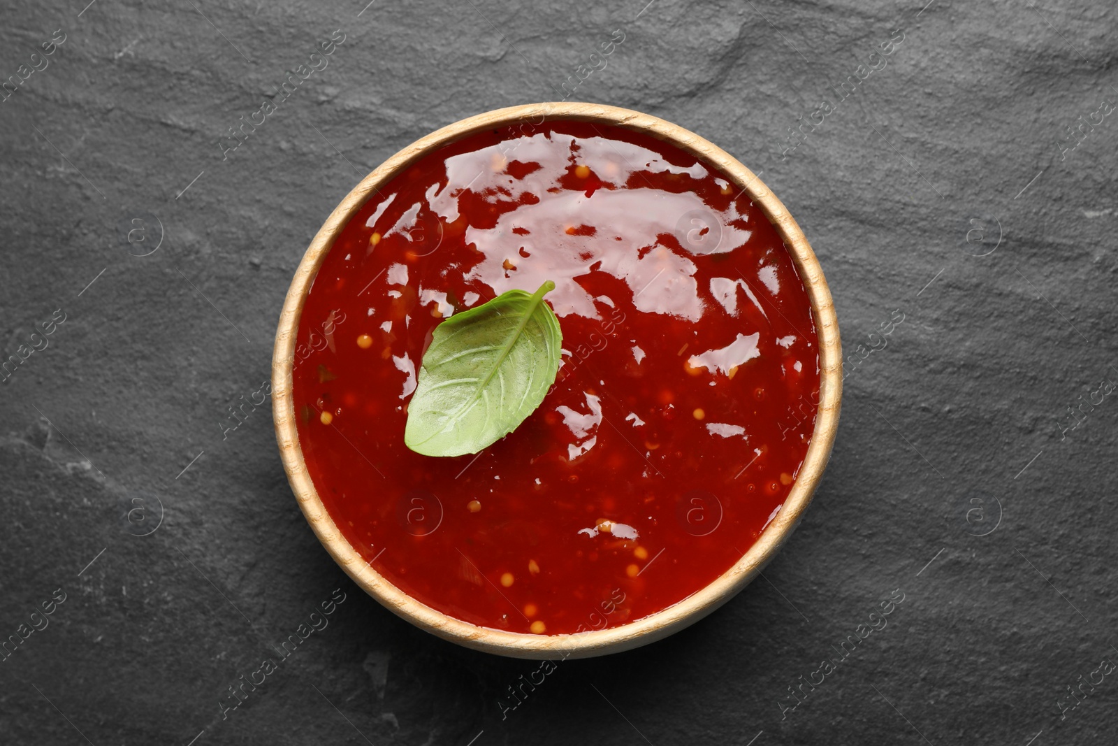 Photo of Spicy chili sauce with basil in bowl on black table, top view