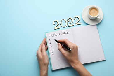 Woman filling list of New Year's resolutions in notebook on light blue background, top view. Space for text