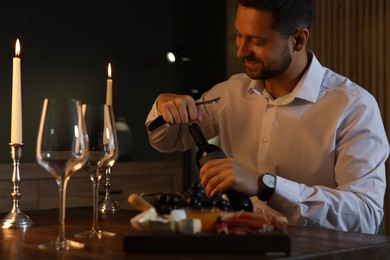 Photo of Romantic dinner. Man opening wine bottle with corkscrew at table indoors