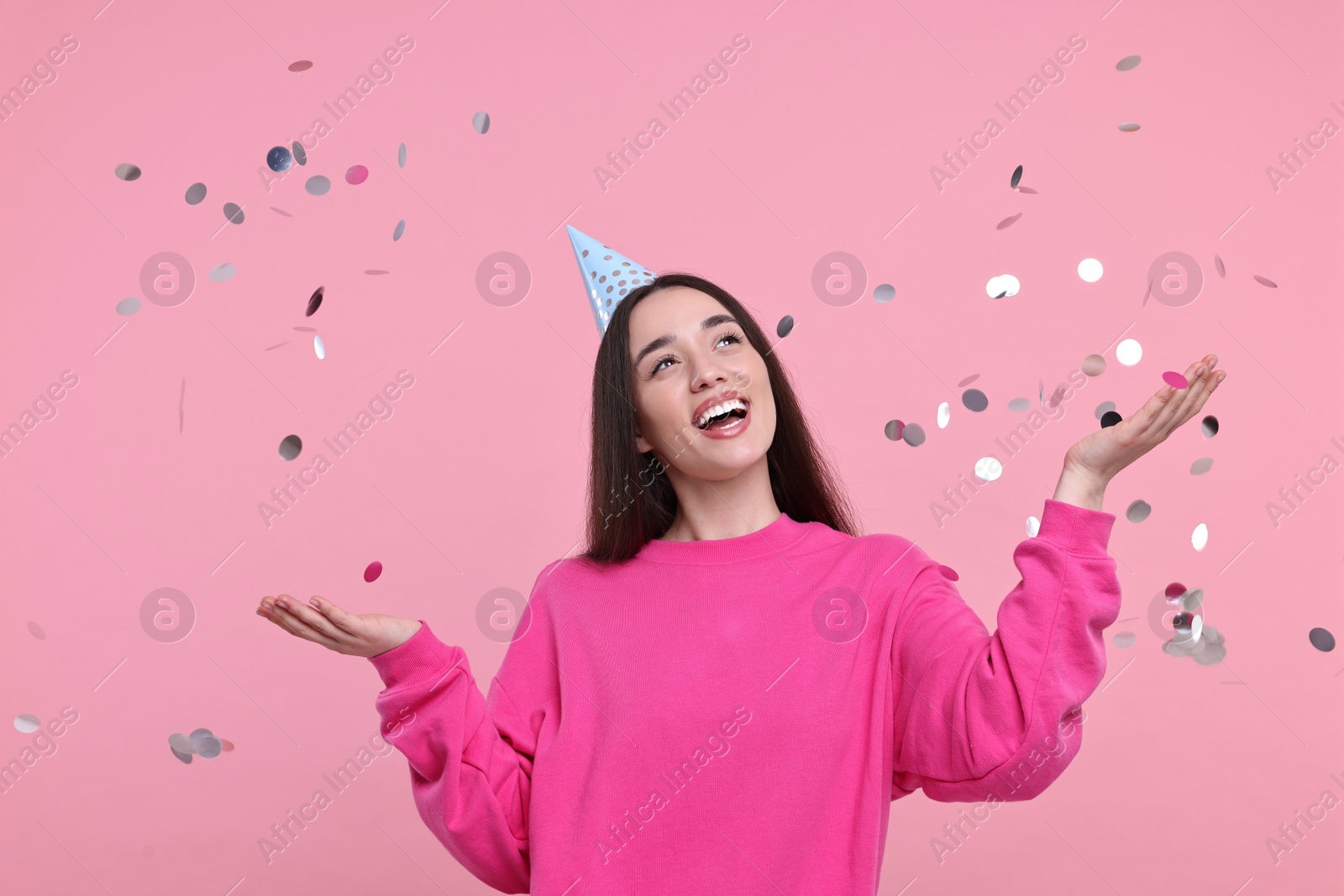 Photo of Happy woman in party hat throwing confetti on pink background