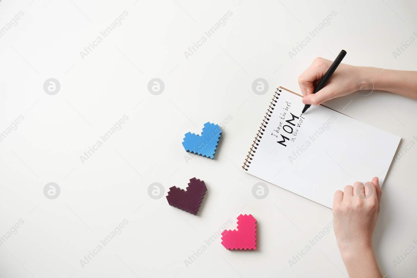 Photo of Little child writing congratulation for Mother's Day in notebook on light background, top view