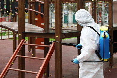 Woman wearing chemical protective suit with disinfectant sprayer on playground. Preventive measure during coronavirus pandemic