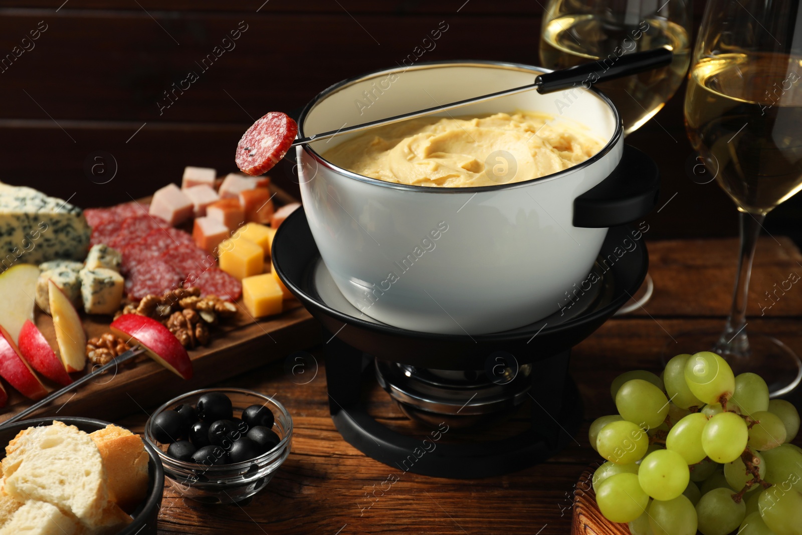 Photo of Fork with piece of sausage, melted cheese in fondue pot and other products on wooden table