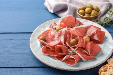 Photo of Slices of tasty cured ham, rosemary and olives on blue wooden table, closeup. Space for text
