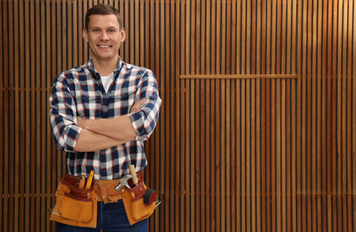 Photo of Handsome carpenter with tool belt near wooden wall. Space for text