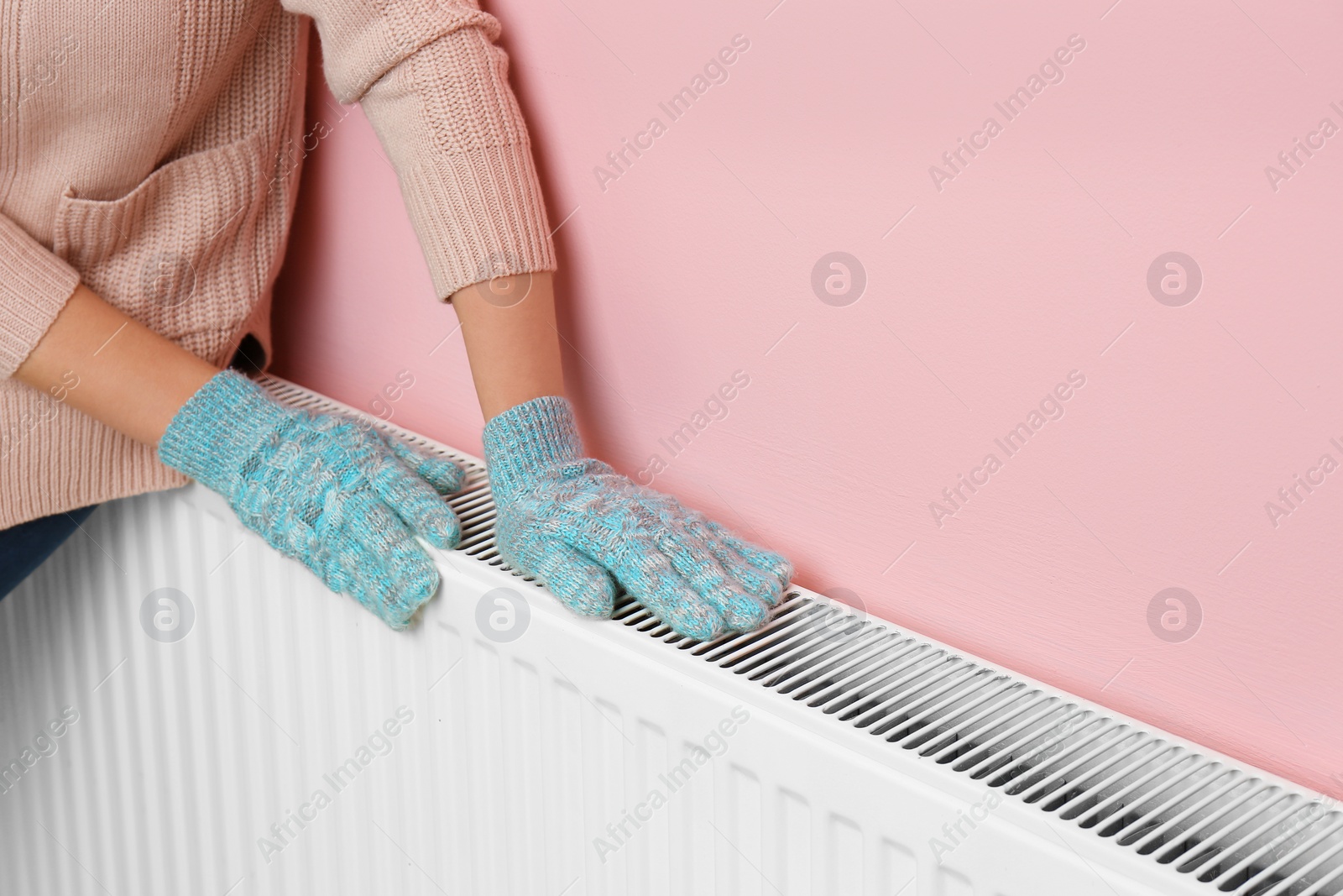 Photo of Woman in gloves warming hands on heating radiator near color wall. Space for text