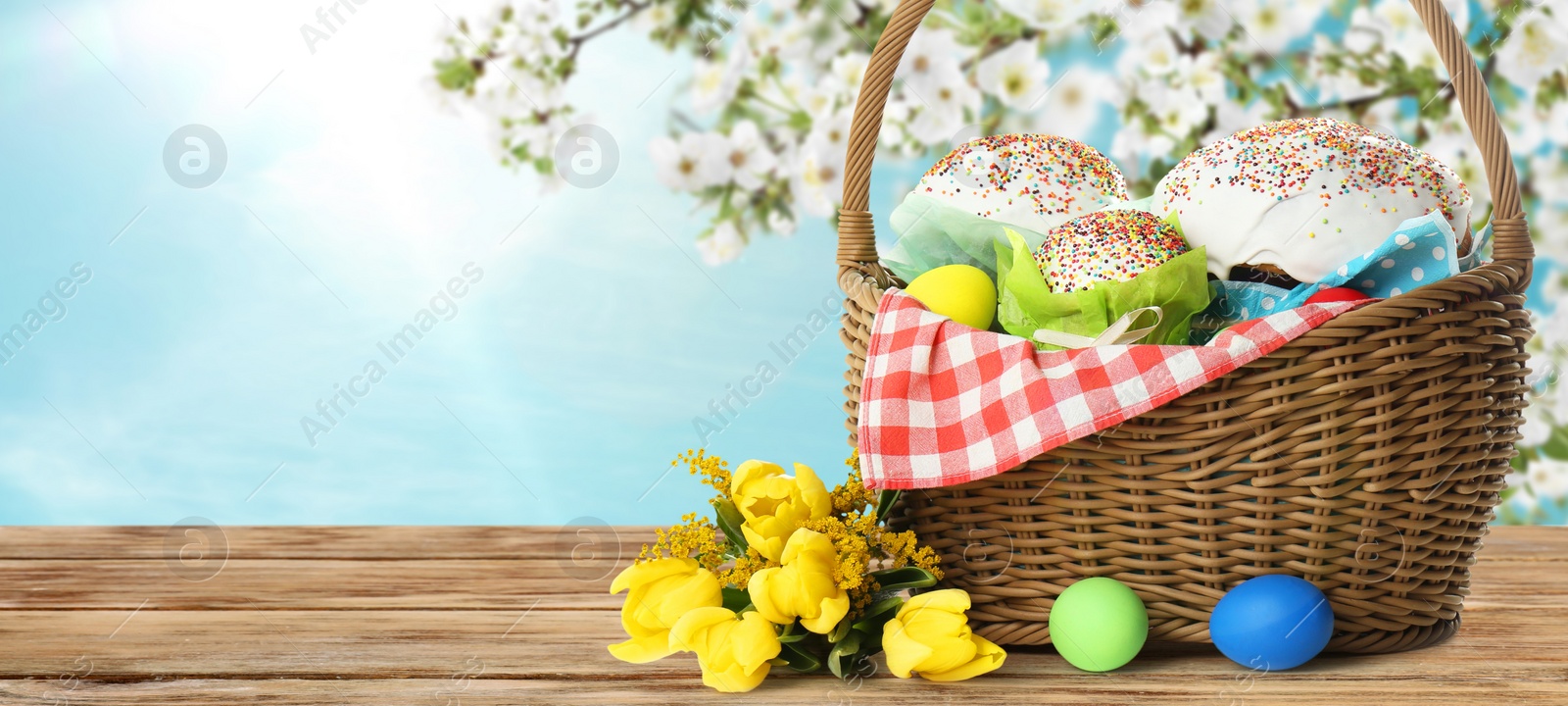 Image of Basket with traditional Easter cake, eggs and flowers on wooden table outdoors, space for text. Banner design