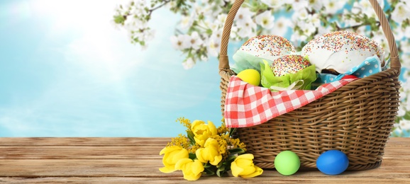 Image of Basket with traditional Easter cake, eggs and flowers on wooden table outdoors, space for text. Banner design