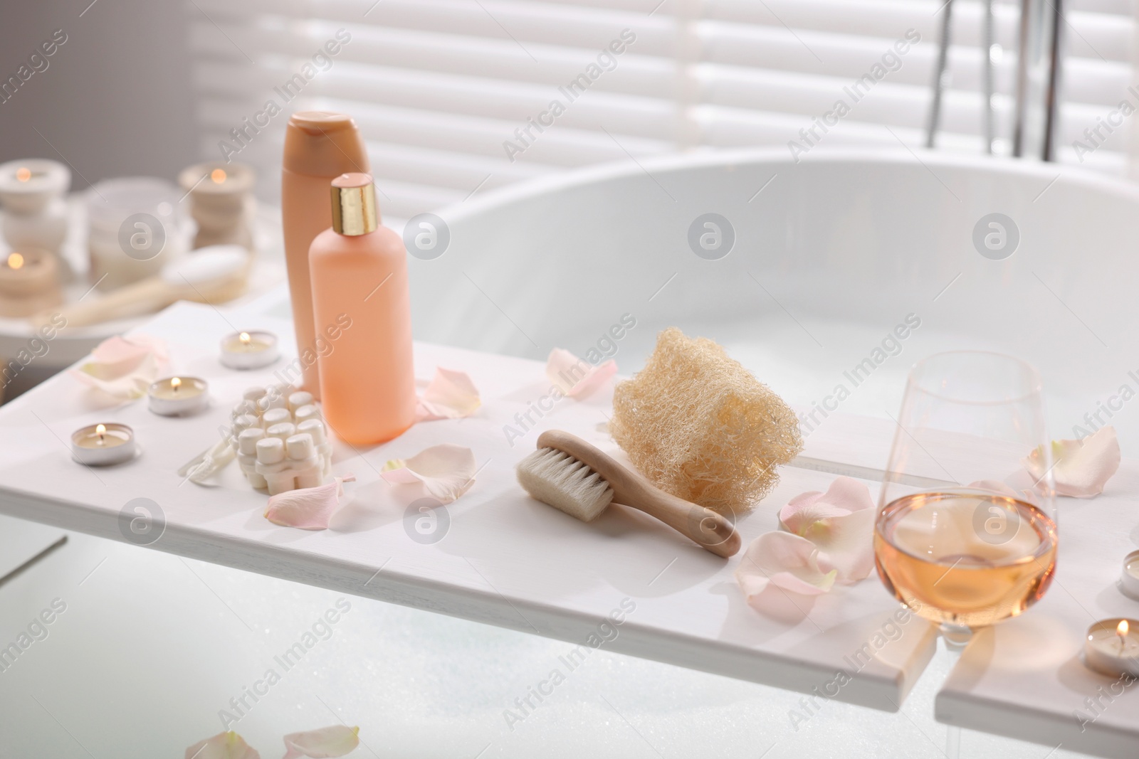 Photo of Wooden tray with wine, toiletries and flower petals on bathtub in bathroom