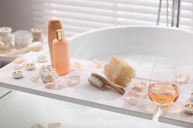 Photo of Wooden tray with wine, toiletries and flower petals on bathtub in bathroom