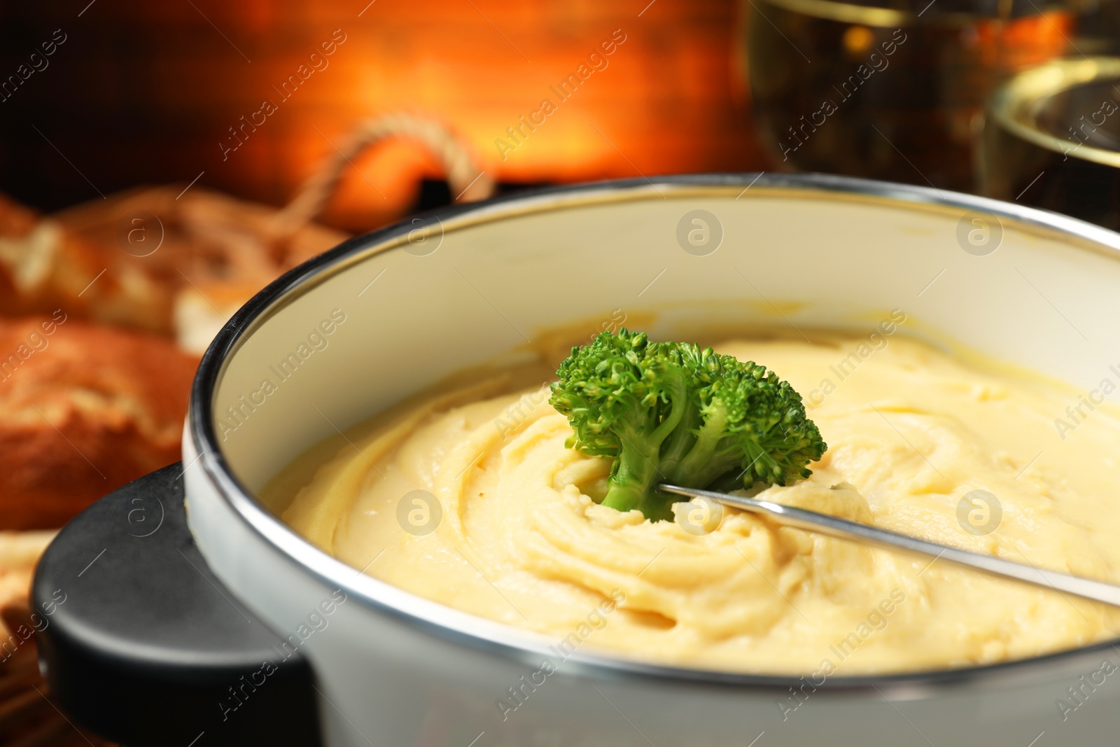 Photo of Dipping piece of broccoli into fondue pot with melted cheese on blurred background, closeup