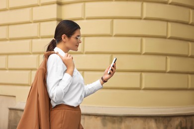 Photo of Young beautiful woman in stylish suit using smartphone near pale yellow wall outdoors