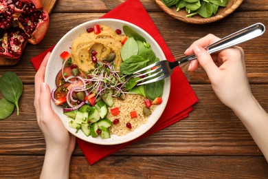 Photo of Woman eating delicious vegan bowl with cucumbers, spinach and bulgur at wooden table, closeup