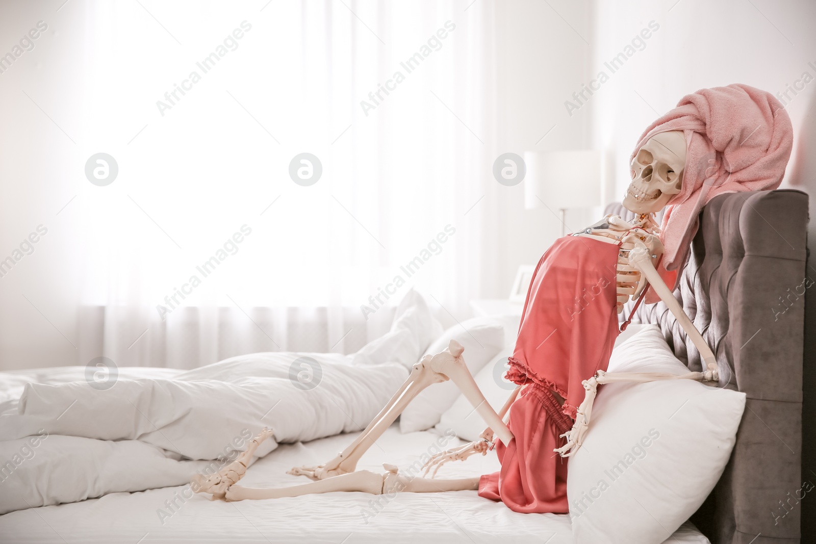 Photo of Human skeleton in silk pajamas and towel sitting on bed indoors