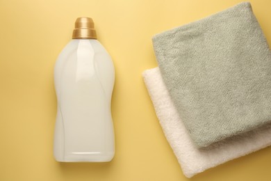 Photo of Bottle of fabric softener and terry towels on pale yellow background, flat lay