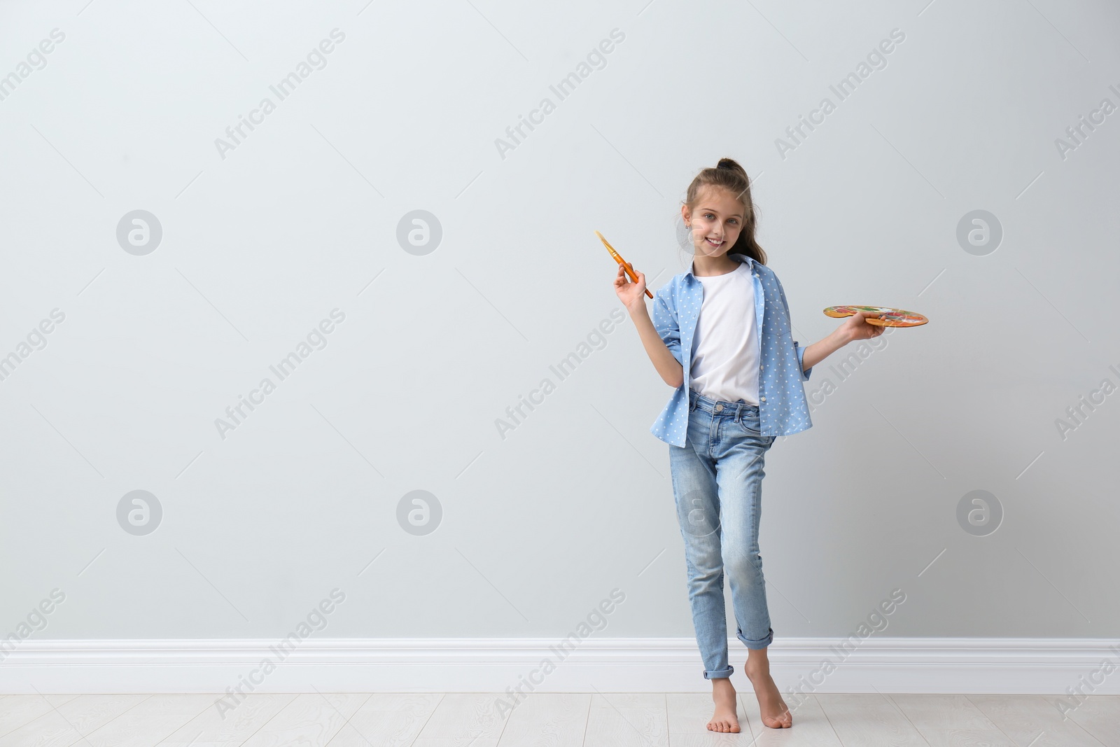 Photo of Little girl with brush and paint palette near light wall indoors. Space for text