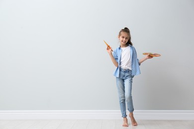 Photo of Little girl with brush and paint palette near light wall indoors. Space for text