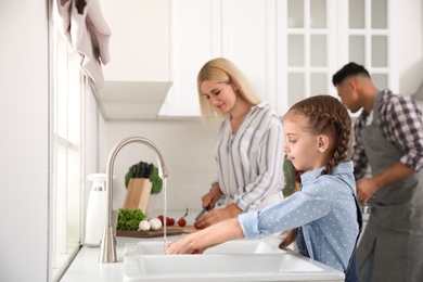 Photo of Little girl washing vegetables and her parents in modern kitchen
