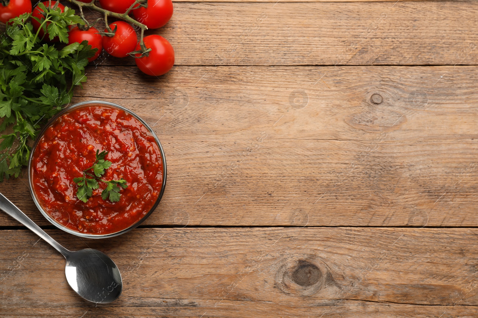 Photo of Delicious adjika sauce in bowl with spoon, parsley and tomatoes on wooden table, flat lay. Space for text