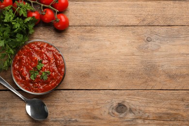 Delicious adjika sauce in bowl with spoon, parsley and tomatoes on wooden table, flat lay. Space for text