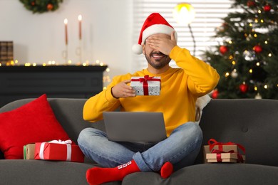 Celebrating Christmas online with exchanged by mail presents. Man in Santa hat covering eyes before opening gift box during video call on laptop at home