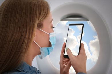 Image of Traveling by airplane during coronavirus pandemic. Woman with face mask taking photo of sky through porthole