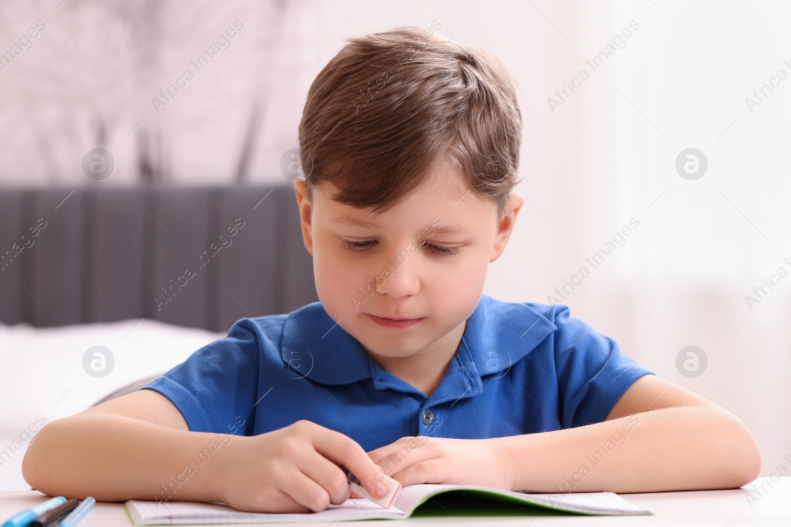 Photo of Little boy erasing mistake in his notebook at white desk indoors