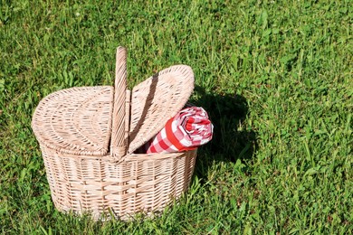 Photo of Rolled checkered tablecloth in picnic basket on green grass outdoors, space for text