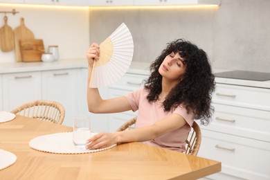 Photo of Young woman waving hand fan to cool herself at table in kitchen