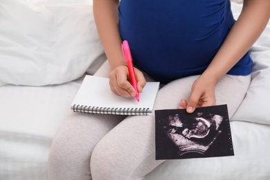 Photo of Pregnant woman with notebook and sonogram choosing name for baby on bed, closeup