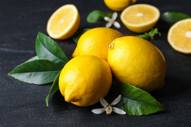 Photo of Many fresh ripe lemons with green leaves and flower on black table, closeup
