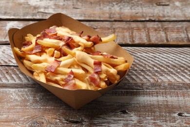 Photo of Tasty potato fries, cheese sauce and bacon in paper container on wooden table, closeup. Space for text