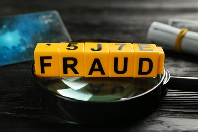 Photo of Word Fraud of yellow cubes with letters and magnifying glass on black wooden background, closeup