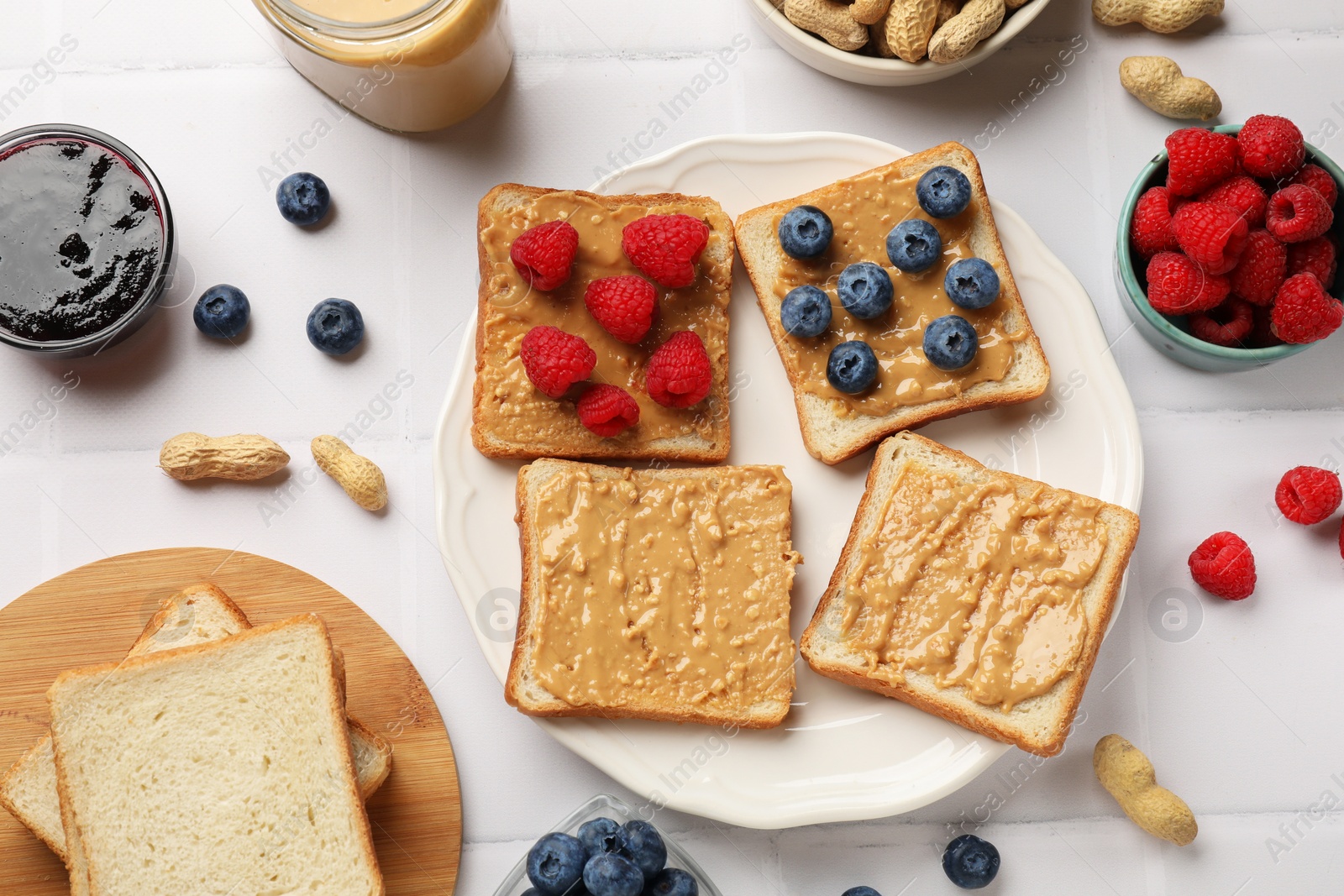 Photo of Delicious toasts with peanut butter, raspberries, blueberries and nuts on white tiled table, flat lay