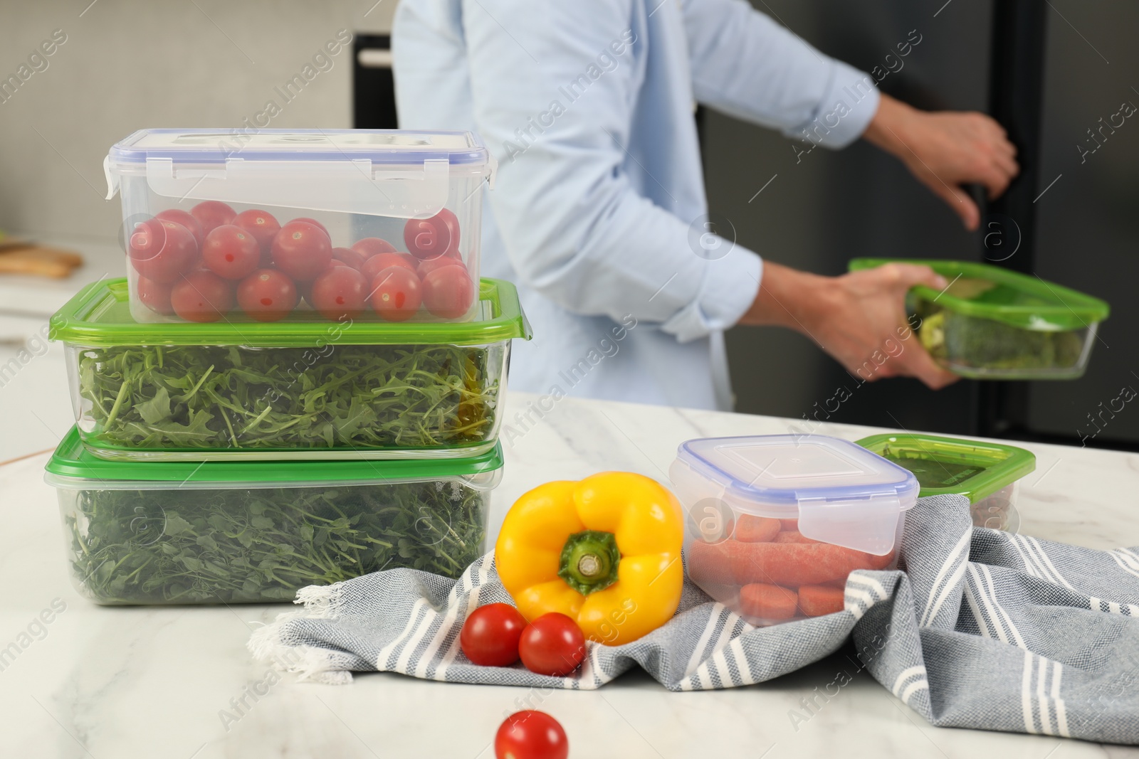 Photo of Glass and plastic containers with different fresh products on white marble table, selective focus. Man near fridge in kitchen