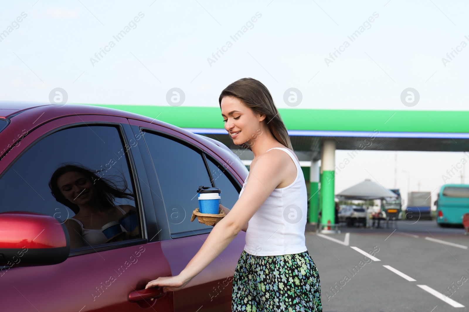 Photo of Beautiful young woman with coffee opening car door at gas station