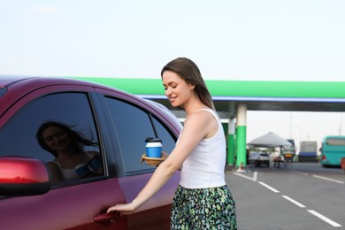 Beautiful young woman with coffee opening car door at gas station