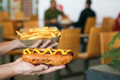 Woman holding fresh tasty hot dog with mustard and french fries in cafe, closeup. Space for text