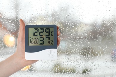 Woman holding digital hygrometer with thermometer near window on rainy day, closeup. Space for text