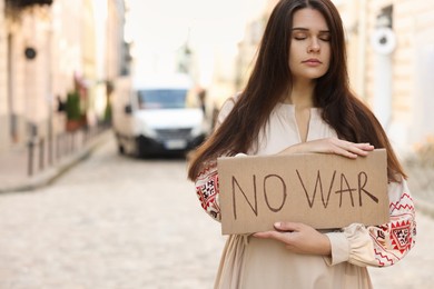Sad woman in embroidered dress holding poster No War on city street. Space for text