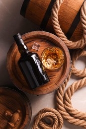 Photo of Whiskey with ice cubes in glass, bottle, wooden barrels and rope on grey marble table, flat lay