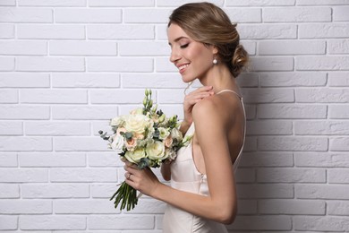 Photo of Young bride with elegant hairstyle holding wedding bouquet near white brick wall