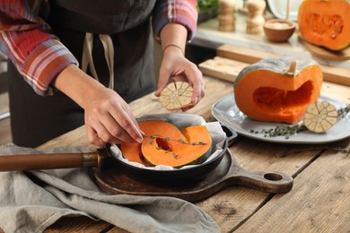 Photo of Woman adding thyme and garlic to pumpkin slices in frying pan at wooden table in kitchen, closeup