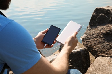 Photo of Man charging mobile phone with power bank on rocky mountain near river, closeup