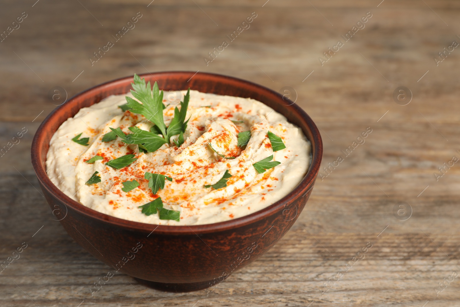 Tasty hummus with parsley and paprika in brown bowl on wooden table Photo of Tasty hummus with parsley and paprika in brown bowl on wooden table