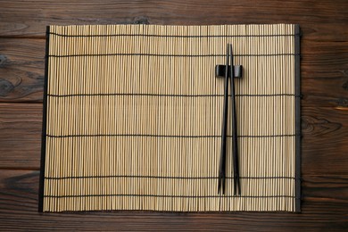 Photo of Bamboo mat with pair of black chopsticks and rest on wooden table, top view