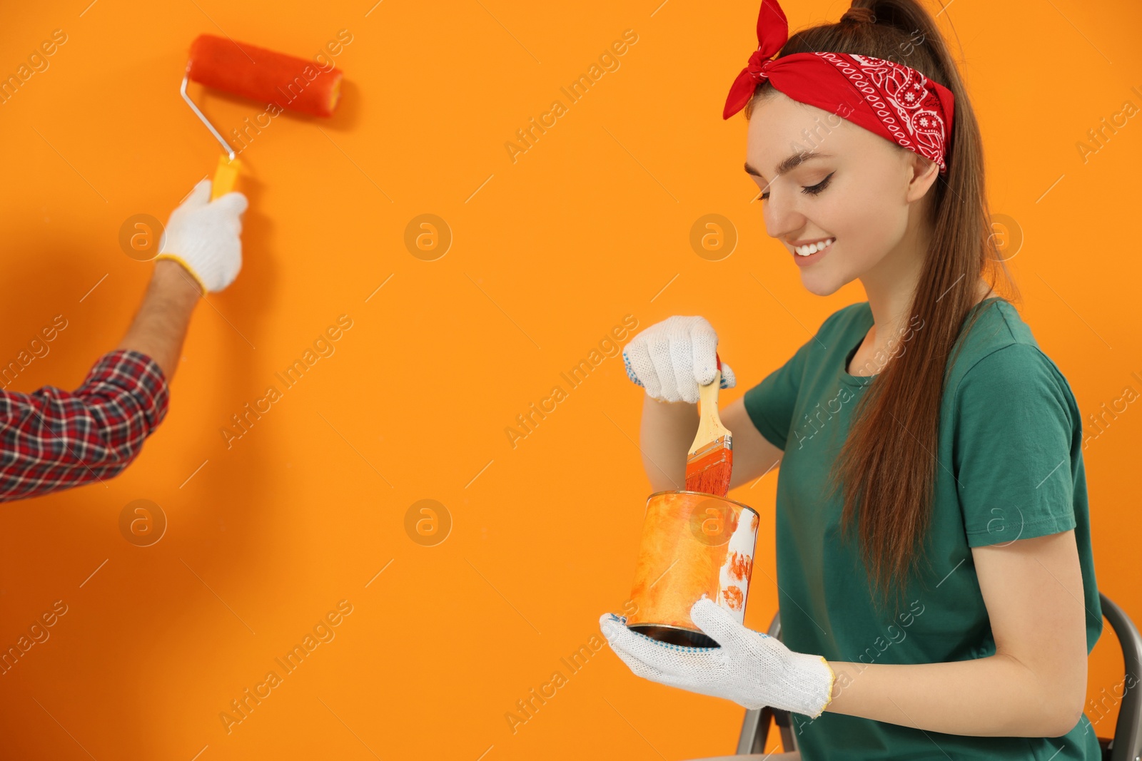 Photo of Man painting orange wall and happy woman holding can of dye with brush. Interior design