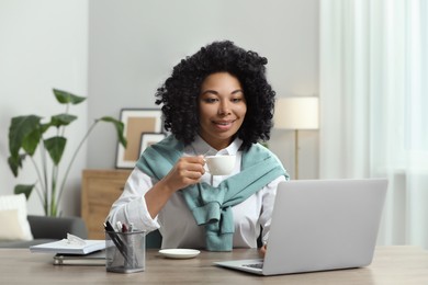 Photo of Happy young woman with cup of drink using laptop at wooden desk indoors