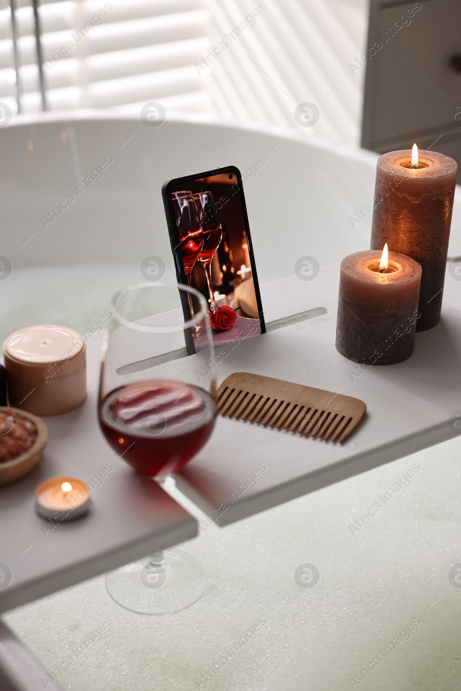 Photo of White wooden tray with smartphone, glass of wine and burning candles on bathtub in bathroom