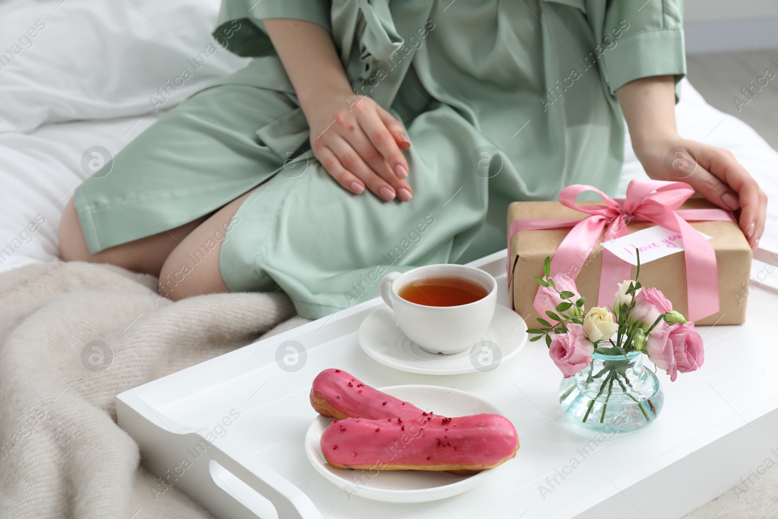 Photo of Tasty breakfast served in bed. Woman with gift box, tea, eclairs, flowers and I Love You card at home, closeup